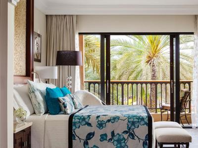 Licht ingerichte hotelkamer met bed, blauw beddengoed en balkon met uitzicht op palmbomen.