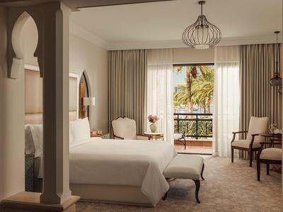 Licht, elegant ingericht hotelkamer met groot bed en balkon met uitzicht op palmbomen.