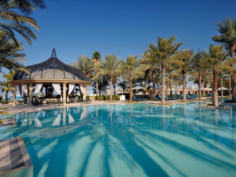 Een rustige zwembadgebied met een prieel en palmbomen onder een heldere blauwe lucht.