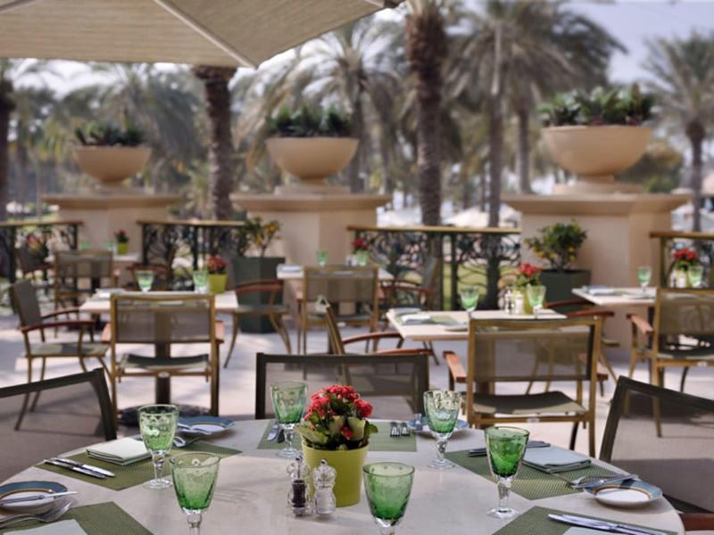 Buitenterras van een restaurant met tafels, groene glazen en bloemen als decoratie bij zonnig weer.