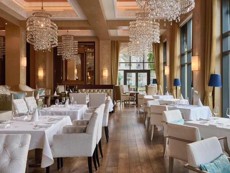 Elegant restaurant met witte tafels, grijze stoelen en kristallen kroonluchters bij daglicht.