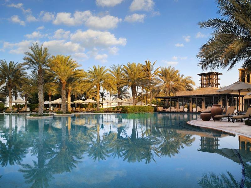 Groot zwembad met palmbomen en ligstoelen voor een resort onder een blauwe lucht