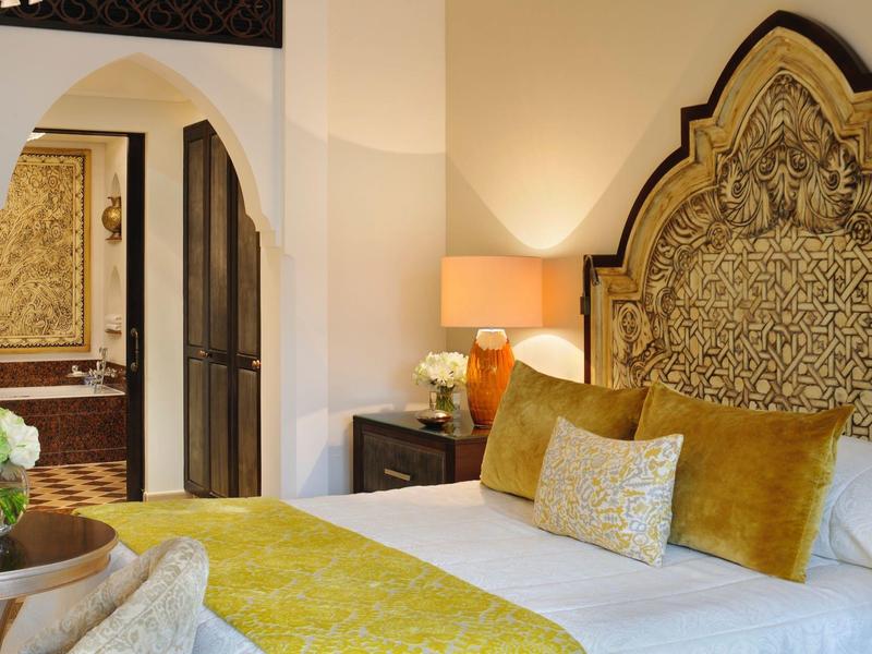 Elegante hotelkamer met groot versierd hoofdeinde, gele kussens en nachtkastje met lamp.