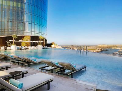 Infinity-zwembad op een hotel dak met ligstoelen en uitzicht op de stad onder een heldere hemel.