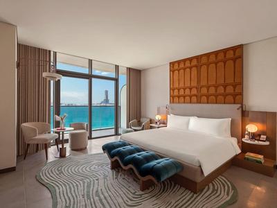 Moderní hotelový pokoj s výhledem na moře, velkou postelí a posezením u oken od podlahy ke stropu.