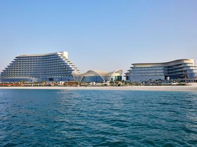 Hotelkomplex mit zwei modernen, terrassenförmigen Gebäuden am Strand vor blauem Meer und klarem Himmel.