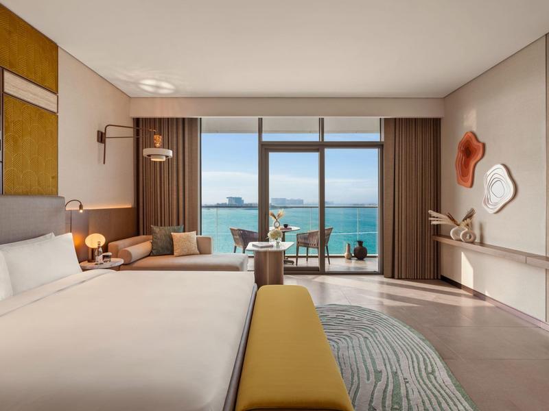 Hotelový pokoj s velkou postelí, posezením a výhledem na moře přes okna od podlahy ke stropu.