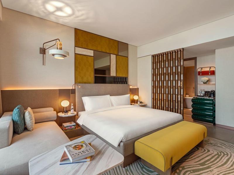 Moderní hotelový pokoj s manželskou postelí, pohovkou, žlutou lavičkou a dekorativní stěnou.