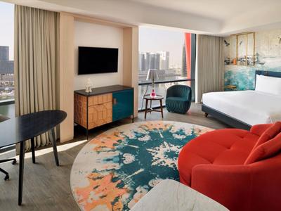 Modernes Hotelzimmer mit großem Bett, buntem Teppich und Stadtblick durch große Fenster.