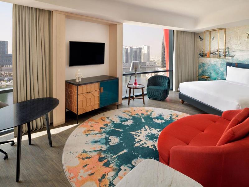 Modernes Hotelzimmer mit großem Bett, buntem Teppich und Stadtblick durch große Fenster.