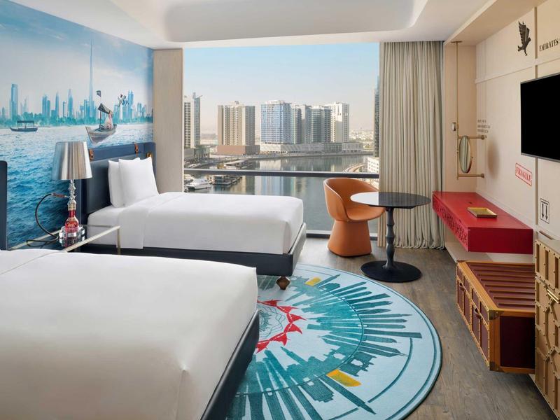 Modernes Hotelzimmer mit zwei Betten, Schreibtisch, Stuhl und Stadtblick durch großes Fenster.