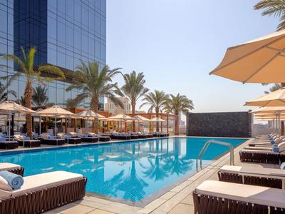 Moderní venkovní bazén s lehátky a slunečníky vedle hotelové budovy a palem.