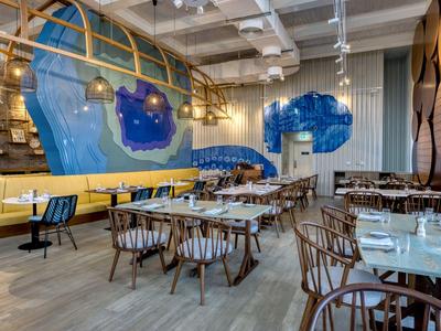 Moderní restaurace s dřevěným nábytkem, žlutou sezením a barevnou nástěnnou malbou.