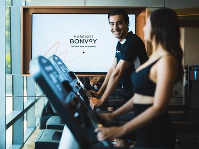 Due persone si allenano su tapis roulant in palestra con uno schermo Marriott Bonvoy sullo sfondo.