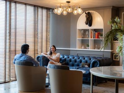 Dva lidé si povídají ve stylovém obývacím pokoji s modrou koženou pohovkou a velkými okny.