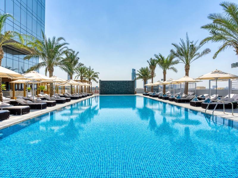 Moderní bazén s lehátky a slunečníky vedle hotelové budovy pod čistou oblohou.