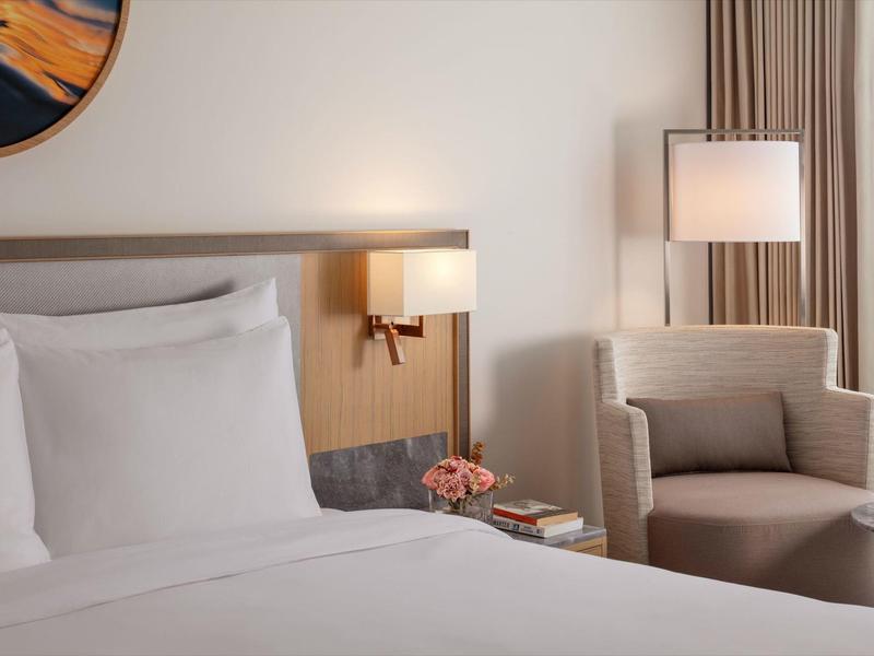 Světý hotelový pokoj s bílou postelí, pohodlným křeslem a lampou u okna.