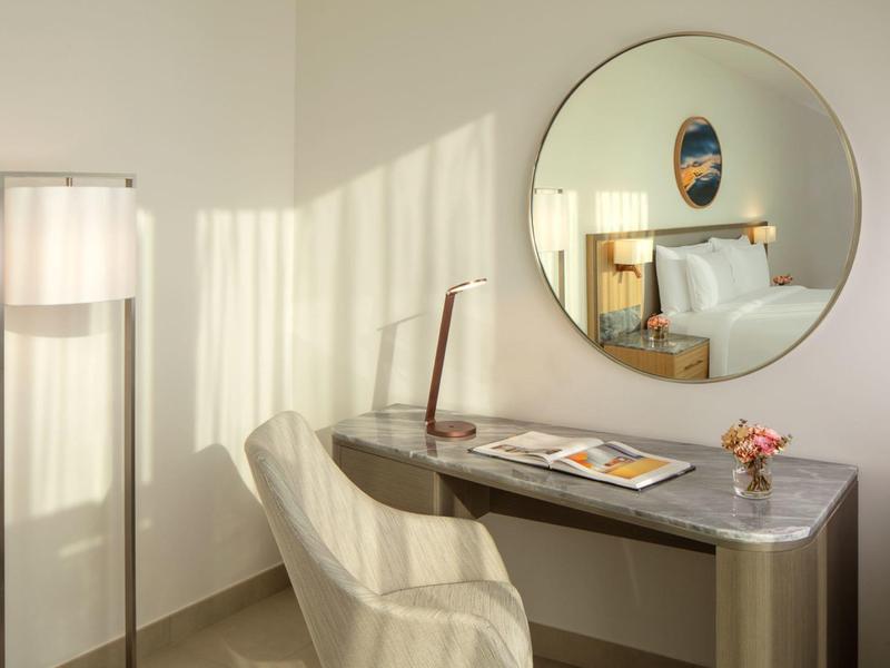Moderní psací stůl se židlí, stojací lampou a kulatým zrcadlem v světlém hotelovém pokoji.