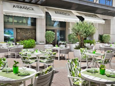 Außenbereich eines Cafés mit runden Tischen, Stühlen und grüner Dekoration bei Tageslicht.