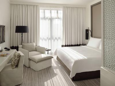 Modern hotel room with double bed, armchair, desk, and large windows with white curtains.