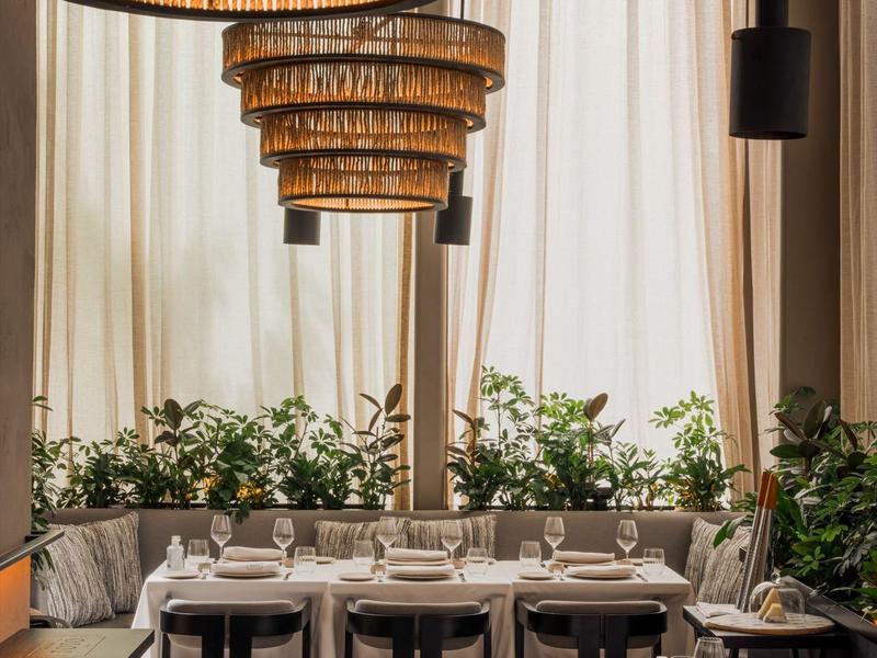 Elegant restaurant with large chandeliers, set tables, and plants in front of light curtains.