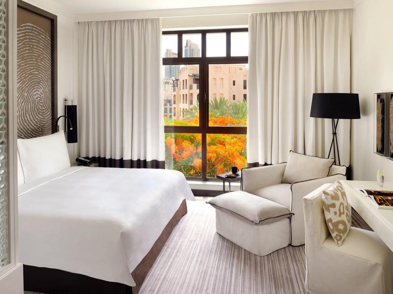 Bright hotel room with large bed, armchairs, desk, and window overlooking city and trees.