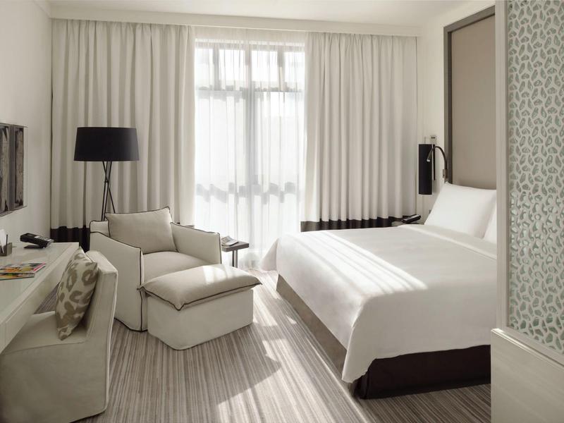 Modern hotel room with double bed, armchair, desk, and large windows with white curtains.