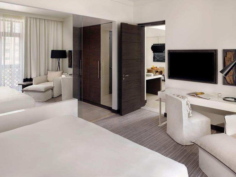 Modern hotel room with two beds, desk, TV, and seating area in daylight.