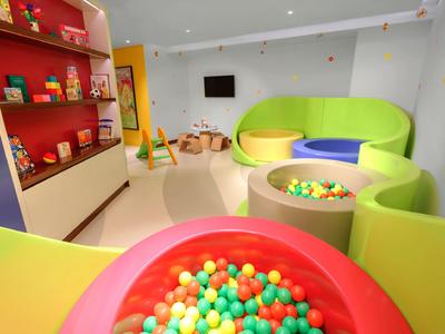 Área de juegos infantil colorida con piscina de bolas, sofás y estanterías en una habitación luminosa.