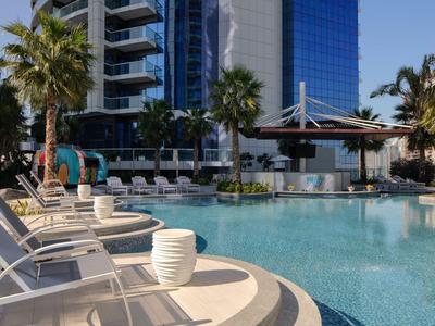 Área de piscina con tumbonas, palmeras y fachada moderna del hotel al fondo.