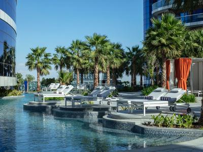 Piscina moderna de hotel con tumbonas y palmeras bajo un cielo azul claro.