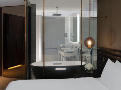 Habitación de hotel moderna con cama blanca, bañera negra y pared de vidrio hacia el baño.
