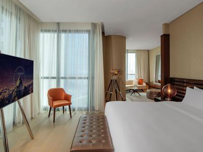 Habitación de hotel moderna con cama grande, televisor en caballete y silla naranja junto a ventanas grandes.