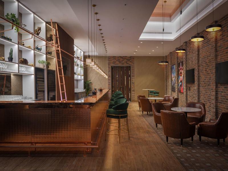 Modern hotel bar area with wooden counter, leather chairs, and warm lighting.