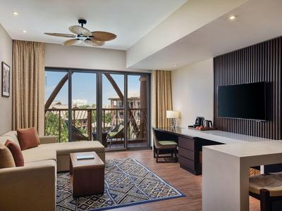 Chambre d'hôtel moderne avec grandes fenêtres, coin salon, bureau et télévision à écran plat.