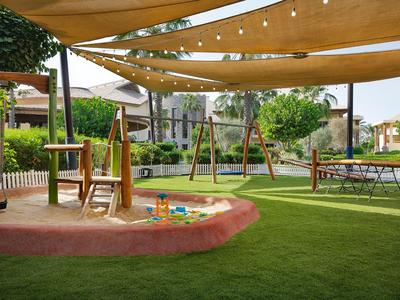 Une aire de jeux ombragée avec bac à sable, balançoires et pelouse dans un jardin d'hôtel.