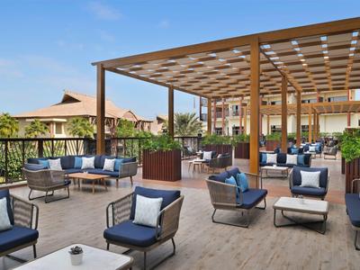 Grande terrasse avec mobilier de lounge et pergola en bois devant des bâtiments d'hôtel sous un ciel clair.