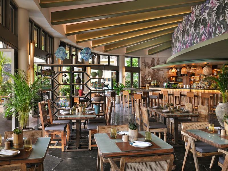 Restaurant lumineux et moderne avec des tables et chaises en bois ainsi que de grandes fenêtres avec des plantes.