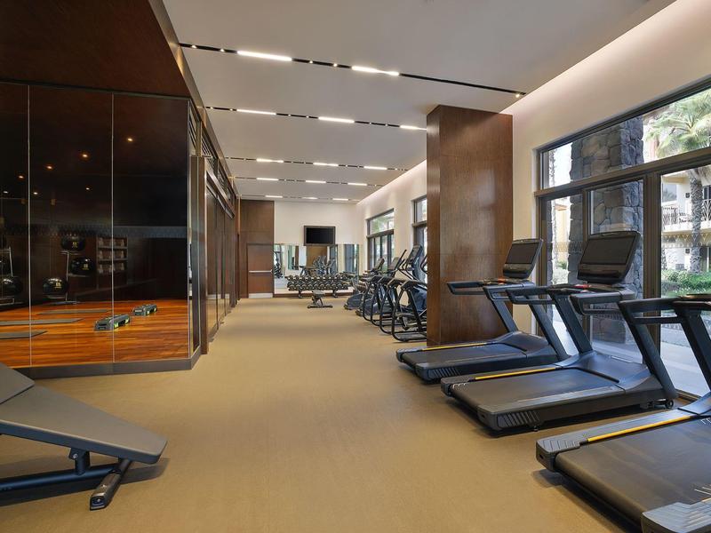 Salle de sport moderne avec tapis de course et équipements, grandes fenêtres et lumière chaleureuse.