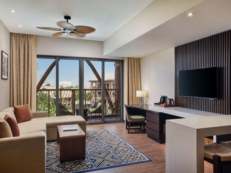 Chambre d'hôtel moderne avec grandes fenêtres, coin salon, bureau et télévision à écran plat.