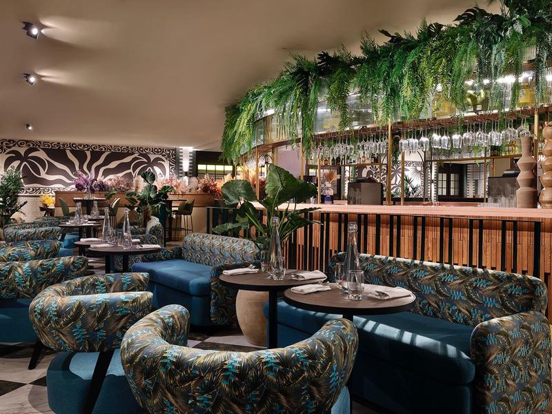 Bar d'hôtel élégante avec chaises à motifs, comptoir en bois et plantes vertes en décoration.