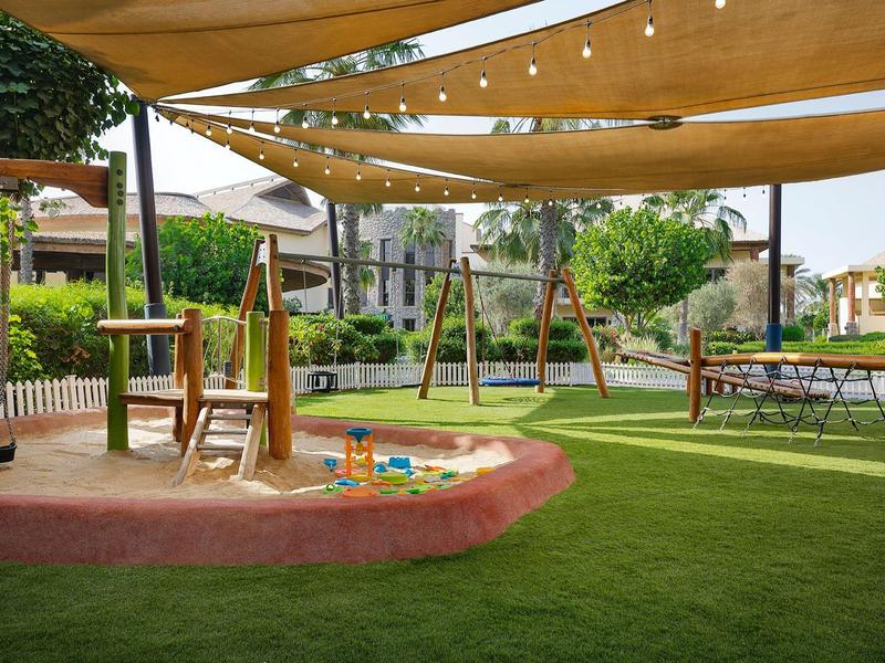 Une aire de jeux ombragée avec bac à sable, balançoires et pelouse dans un jardin d'hôtel.