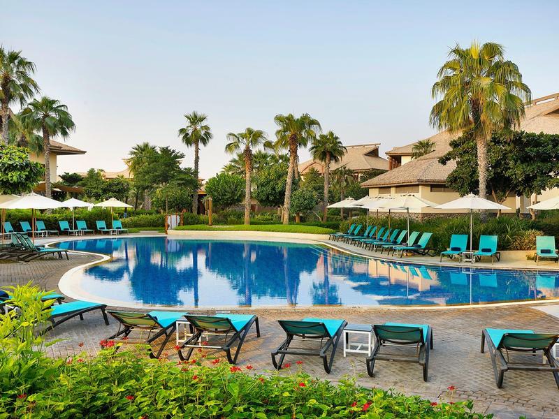 Zone de piscine avec chaises longues et palmiers dans un complexe hôtelier sous un ciel dégagé.