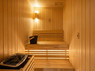 Světlá dřevěná sauna s lavicemi, pásovým osvětlením a saunovým kamenem s kbelíky.