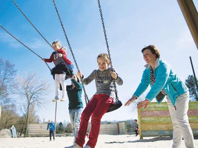 Drei Personen schaukeln auf einem Spielplatz bei blauem Himmel.
