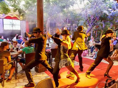 Tanzende Menschen auf einer Bühne vor einem sitzenden Publikum in einer Außenumgebung.