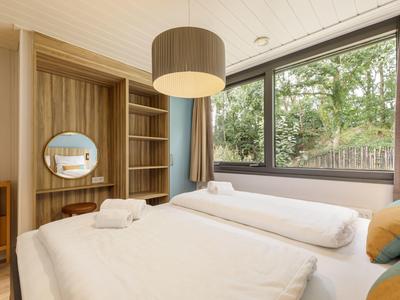 Moderní hotelový pokoj s dvojlůžkem, závěsnou lampou a velkým oknem s výhledem na zeleň.