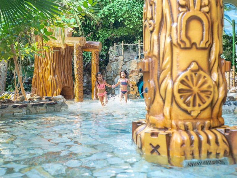 Děti si hrají v tropickém vnitřním bazénu s dekorativními sloupy a vodními fontánami.