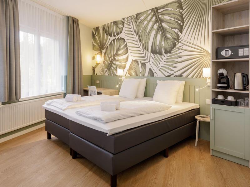 Moderní hotelový pokoj s velkou postelí, lampami, tapetou s listy a jasným denním světlem z okna.