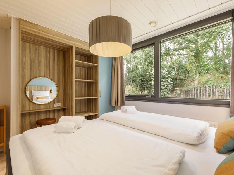 Moderní hotelový pokoj s dvojlůžkem, závěsnou lampou a velkým oknem s výhledem na zeleň.